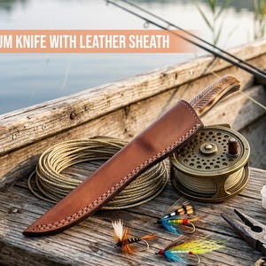 Pu&ograve; includere: Un coltello con manico in legno decorato all'interno di un fodero in pelle marrone, posizionato accanto a un mulinello da pesca, una bobina di filo, mosche da pesca colorate e pinze in metallo su una superficie in legno con il testo PREMIUM KNIFE WITH LEATHER SHEATH.