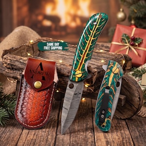 May include: A folding knife with a Damascus steel blade and a green and yellow patterned handle, accompanied by a matching folded knife and a brown leather sheath embossed with the name Andrew and a brass snap closure button.