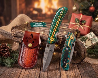 May include: A folding knife with a Damascus steel blade and a green and yellow patterned handle, accompanied by a matching folded knife and a brown leather sheath embossed with the name Andrew and a brass snap closure button.
