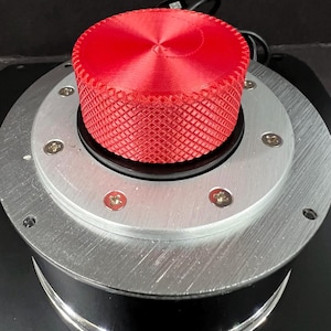 May include: A close-up of a red, textured knob with a crosshatch pattern, mounted on a silver metal base. The knob has a ridged top and is secured with small screws. The base sits on a black surface.