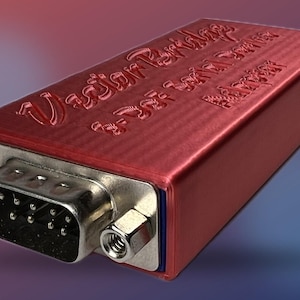 May include: A red rectangular electronic device with a silver connector and text that reads "Vector Peripherals 9-Pin Serial Adapter". The device has a metallic finish and is set against a gradient background.
