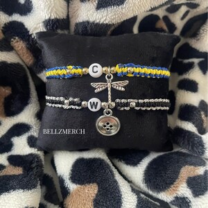 May include: Two macrame bracelets with letter beads and charms. One bracelet is blue and yellow, the other is black and silver. A dragonfly charm and a button charm hang from the bracelets. The bracelets are displayed on a black pillow.