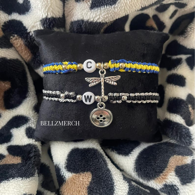 May include: Two macrame bracelets with letter beads and charms. One bracelet is blue and yellow, the other is black and silver. A dragonfly charm and a button charm hang from the bracelets. The bracelets are displayed on a black pillow.