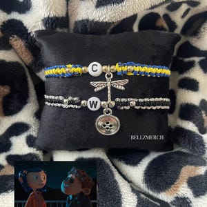May include: Two macrame bracelets with letter charms and dragonfly and button pendants. One bracelet is blue and yellow, the other is black and white. The bracelets are displayed on a black surface with a leopard print background. The text "BELLZMERCH" is visible.