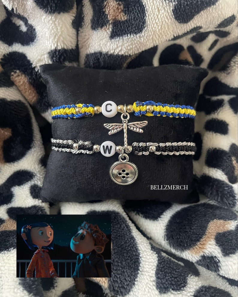 May include: Two macrame bracelets with letter charms and dragonfly and button pendants. One bracelet is blue and yellow, the other is black and white. The bracelets are displayed on a black surface with a leopard print background. The text "BELLZMERCH" is visible.