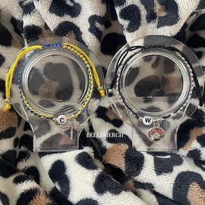 May include: Two bracelets displayed on clear stands. One bracelet is yellow and blue, with a dragonfly charm and the letter "C". The other is black and silver, with a charm and the letter "W". The background is a leopard print.