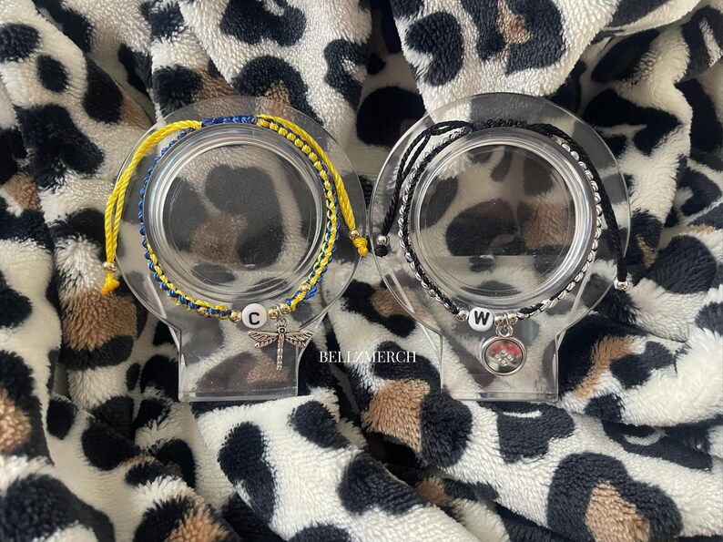 May include: Two bracelets displayed on clear stands. One bracelet is yellow and blue, with a dragonfly charm and the letter "C". The other is black and silver, with a charm and the letter "W". The background is a leopard print.