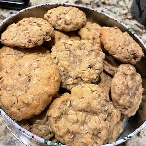 Puede incluir: Una lata llena de varias galletas de avena de color marrón dorado. Las galletas son redondas y tienen avena visible en la superficie. La lata tiene un borde blanco con un patrón rojo y verde.