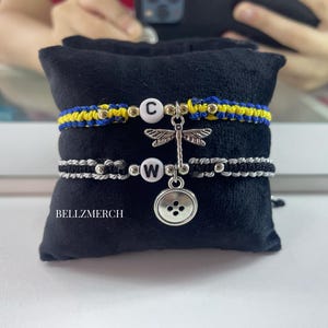 May include: Two macrame bracelets with letter charms and decorative charms. One bracelet is blue and yellow, the other is black and silver. A dragonfly charm and a button charm hang between the bracelets. The text "BELLZMERCH" is visible.