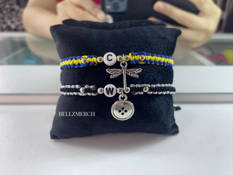 May include: Two macrame bracelets with letter charms and decorative charms. One bracelet is blue and yellow, the other is black and silver. A dragonfly charm and a button charm hang between the bracelets. The text "BELLZMERCH" is visible.