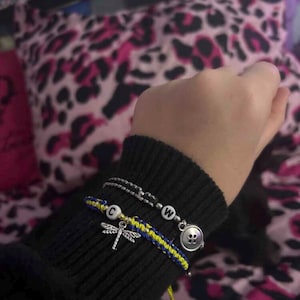 May include: A black knit sleeve displays three bracelets. One is a silver chain with a white letter bead. Another is a woven blue and yellow bracelet with a dragonfly charm and a white letter bead. The third bracelet has a button charm.