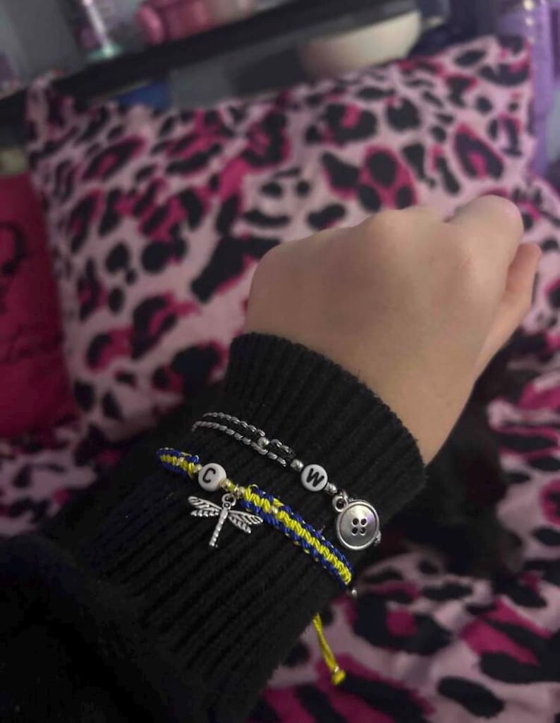 May include: A black knit sleeve displays three bracelets. One is a silver chain with a white letter bead. Another is a woven blue and yellow bracelet with a dragonfly charm and a white letter bead. The third bracelet has a button charm.