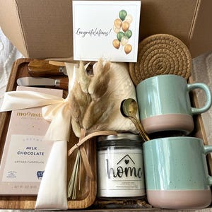 May include: A gift box containing a Moonstruck milk chocolate bar, two speckled light green and pink mugs, a glass candle jar labeled home, a wooden tray, two metal spreaders, a round woven coaster, a small dried floral bouquet, and a card.