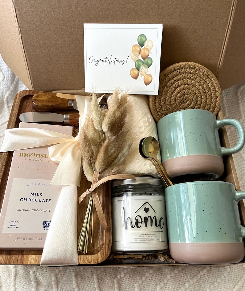 May include: A gift box containing a Moonstruck milk chocolate bar, two speckled light green and pink mugs, a glass candle jar labeled home, a wooden tray, two metal spreaders, a round woven coaster, a small dried floral bouquet, and a card.