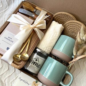 May include: A gift box containing a wooden serving board with two gold spoons and a cream ribbon, a milk chocolate bar, two speckled light green mugs with tan bases, a candle labeled home, two woven coasters, a white cloth, and a congratulations card.