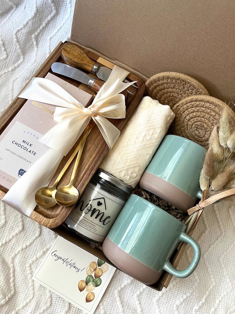 May include: A gift box containing a wooden serving board with two gold spoons and a cream ribbon, a milk chocolate bar, two speckled light green mugs with tan bases, a candle labeled home, two woven coasters, a white cloth, and a congratulations card.