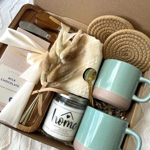 May include: A gift box containing two speckled light green and tan mugs, a glass candle jar labeled home, a wooden tray, a chocolate bar, two round woven coasters, a small gold spoon, a metal spreader, and a bundle of dried decorative grasses.