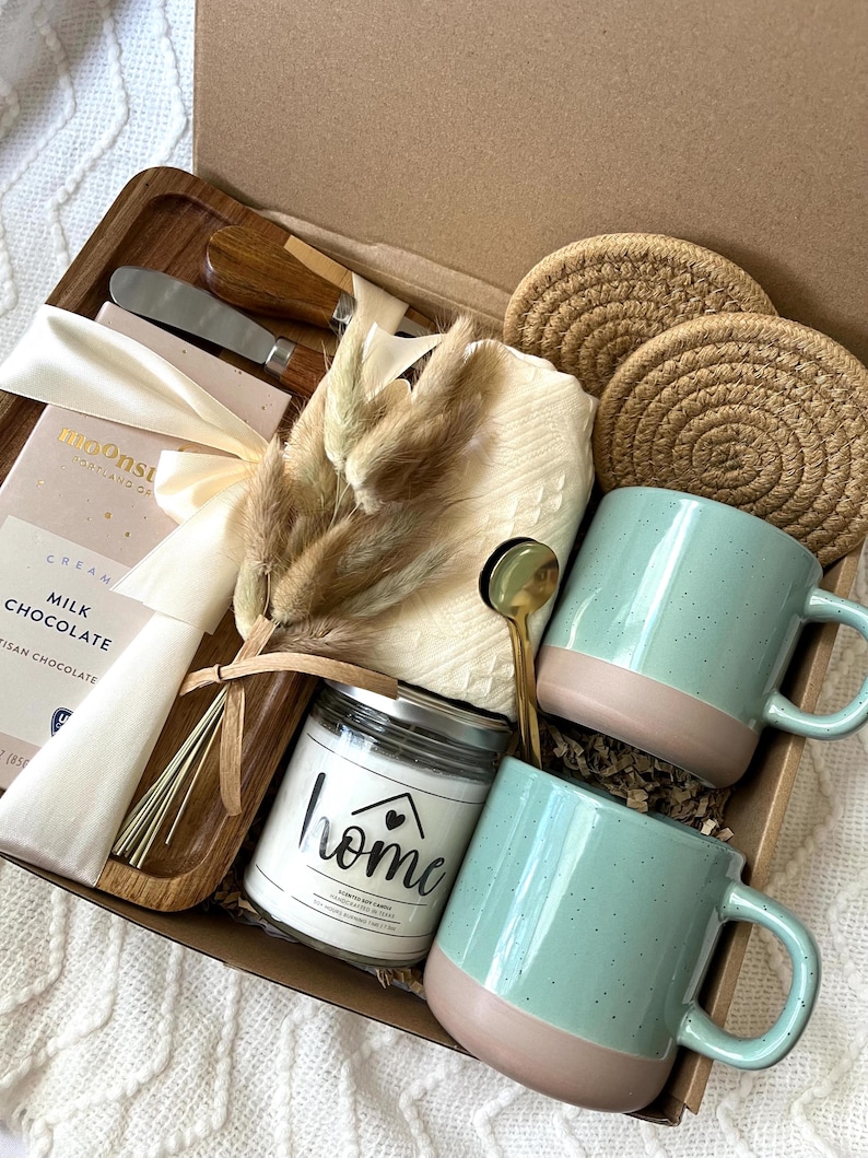 May include: A gift box containing two speckled light green and tan mugs, a glass candle jar labeled home, a wooden tray, a chocolate bar, two round woven coasters, a small gold spoon, a metal spreader, and a bundle of dried decorative grasses.