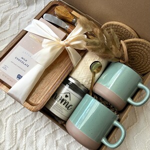 May include: A gift box containing a wooden tray with a milk chocolate bar tied with a cream ribbon, two metal spreaders with wooden handles, two speckled light green and tan mugs, a candle labeled home, a gold spoon, and two round woven coasters.
