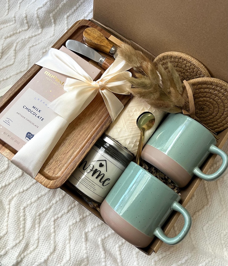 May include: A gift box containing a wooden tray with a milk chocolate bar tied with a cream ribbon, two metal spreaders with wooden handles, two speckled light green and tan mugs, a candle labeled home, a gold spoon, and two round woven coasters.