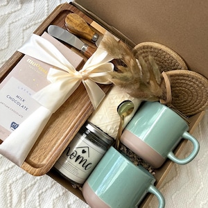 May include: A gift box containing a wooden serving tray, a milk chocolate bar with a white ribbon, two metal spreaders with wooden handles, a candle labeled home, two speckled sage green and tan mugs, a gold spoon, and two round woven coasters.