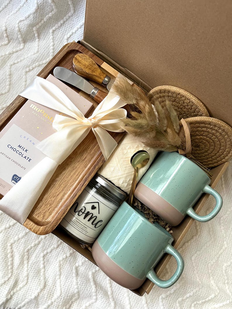 May include: A gift box containing a wooden serving tray, a milk chocolate bar with a white ribbon, two metal spreaders with wooden handles, a candle labeled home, two speckled sage green and tan mugs, a gold spoon, and two round woven coasters.