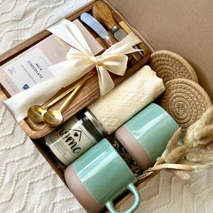 May include: A gift box containing a wooden tray with a bar of milk chocolate, two gold spoons, and two cheese knives. Included are two speckled mint green and tan mugs, a candle labeled home, a folded cream textile, and two round jute coasters.