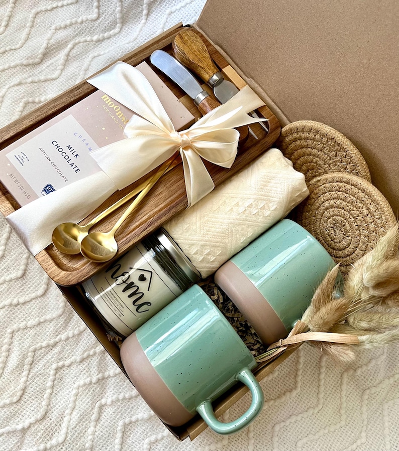 May include: A gift box containing a wooden tray with a bar of milk chocolate, two gold spoons, and two cheese knives. Included are two speckled mint green and tan mugs, a candle labeled home, a folded cream textile, and two round jute coasters.