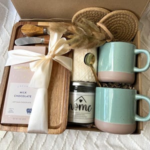 May include: A gift box containing a wooden serving tray, a 3 oz milk chocolate bar, two speckled light green and tan mugs, a small glass candle jar labeled home, two round woven coasters, a gold spoon, a cheese knife, and dried grass.