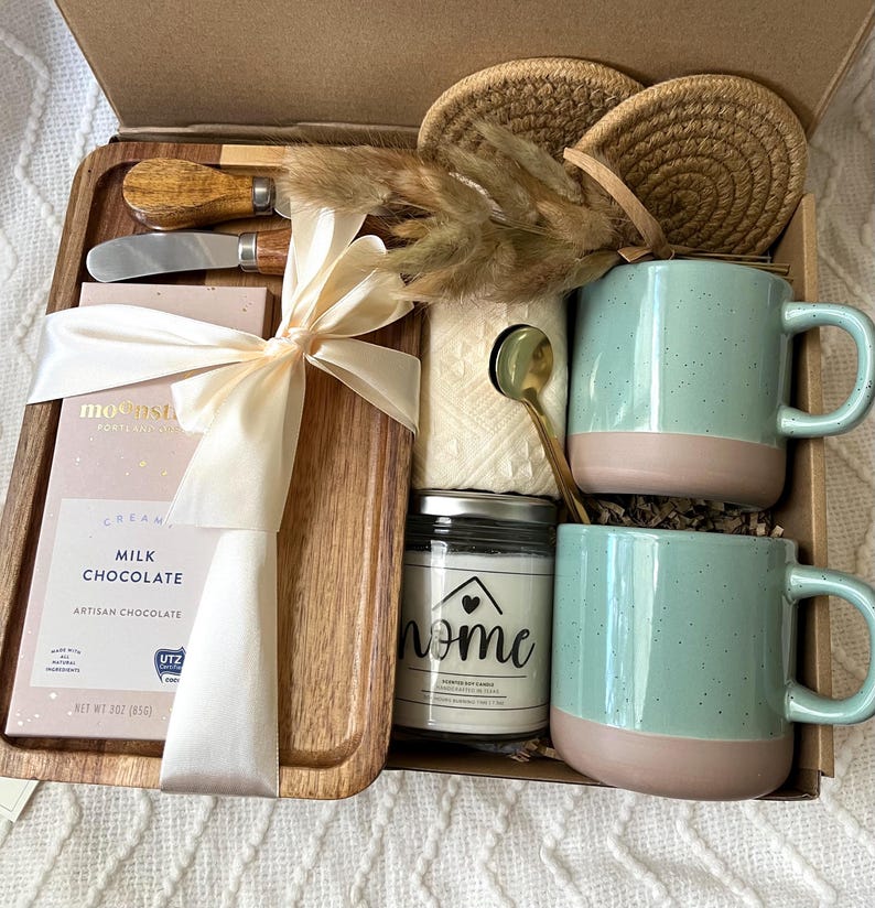 May include: A gift box containing a wooden serving tray, a 3 oz milk chocolate bar, two speckled light green and tan mugs, a small glass candle jar labeled home, two round woven coasters, a gold spoon, a cheese knife, and dried grass.