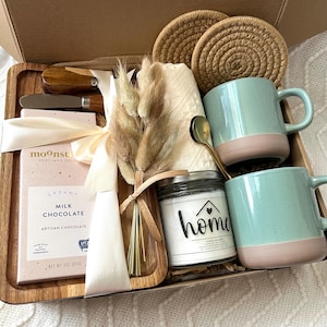 May include: A gift box containing a wooden tray, two speckled light blue and pink mugs, a gold spoon, a candle labeled home, a Moonstruck milk chocolate bar, two woven coasters, a small bouquet of dried grass, and a wooden handled spreader.