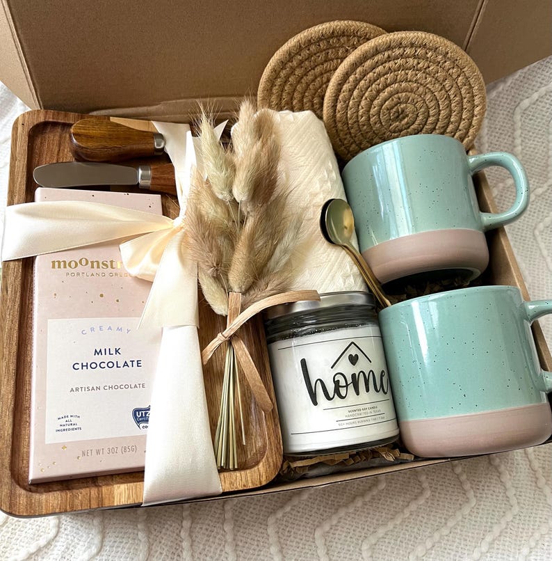 May include: A gift box containing a wooden tray, two speckled light blue and pink mugs, a gold spoon, a candle labeled home, a Moonstruck milk chocolate bar, two woven coasters, a small bouquet of dried grass, and a wooden handled spreader.