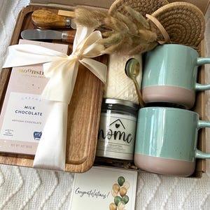 May include: A gift box containing a wooden serving board, a Moonstruck milk chocolate bar, two speckled light green and tan mugs, a candle labeled home, two woven coasters, a gold spoon, a cheese spreader, dried grass, and a Congratulations card.