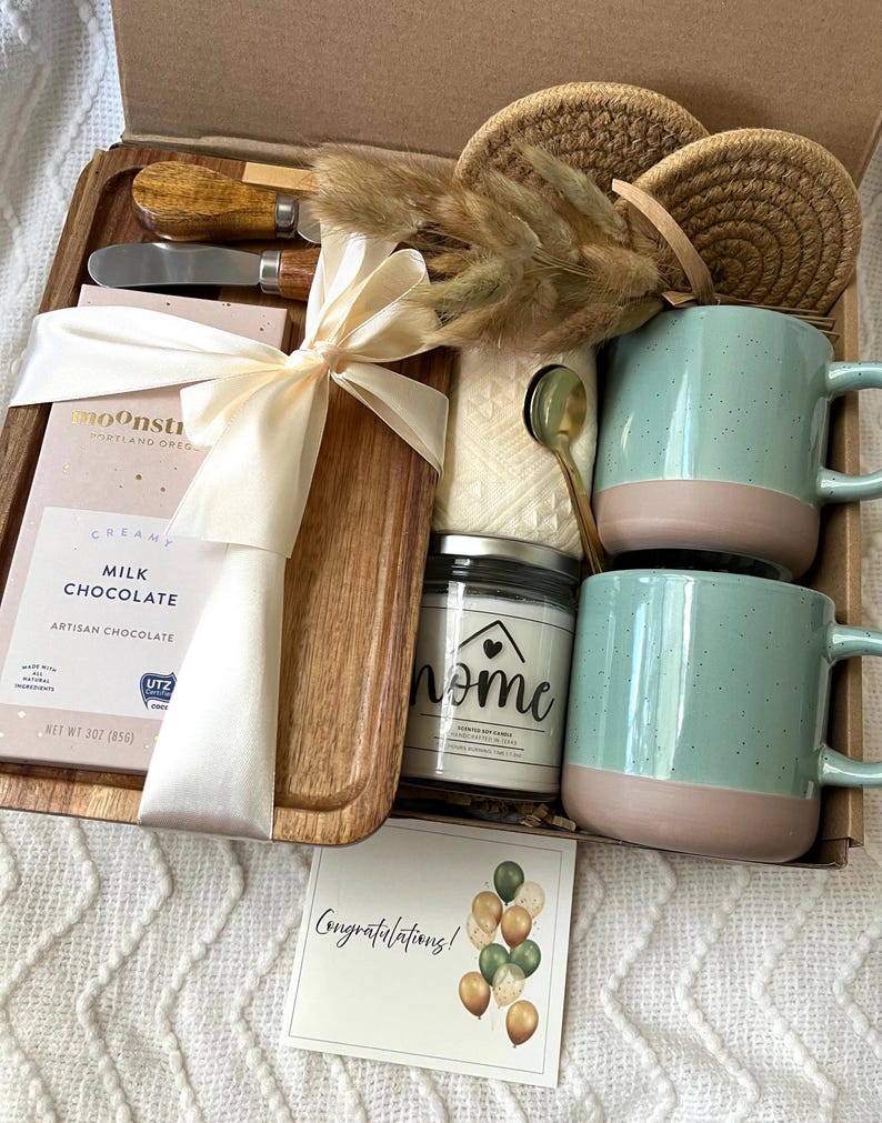 May include: A gift box containing a wooden serving board, a Moonstruck milk chocolate bar, two speckled light green and tan mugs, a candle labeled home, two woven coasters, a gold spoon, a cheese spreader, dried grass, and a Congratulations card.