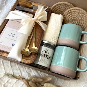 May include: A gift box containing a wooden serving board, two gold spoons, two cheese knives with wooden handles, a bar of Moonstruck milk chocolate, two round jute coasters, a white textured cloth, a candle labeled home, and dried grass.