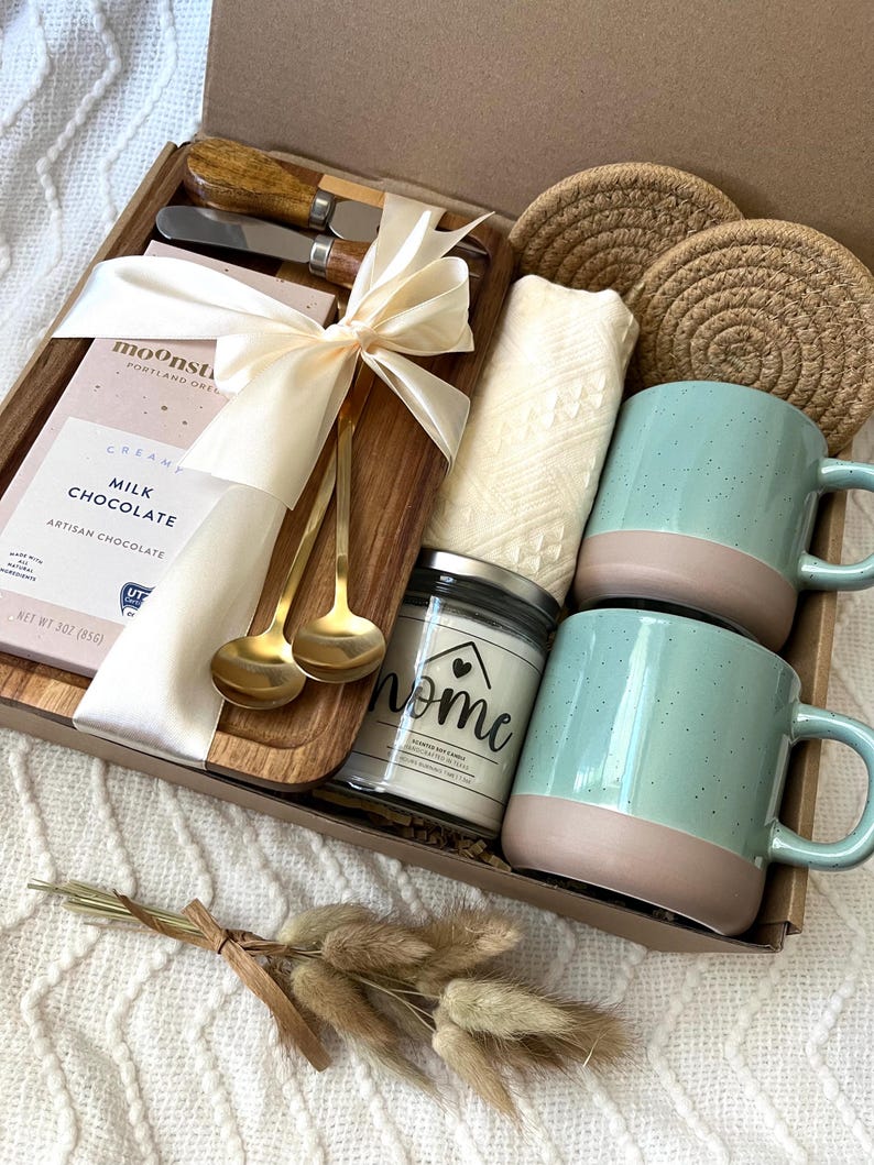 May include: A gift box containing a wooden serving board, two gold spoons, two cheese knives with wooden handles, a bar of Moonstruck milk chocolate, two round jute coasters, a white textured cloth, a candle labeled home, and dried grass.