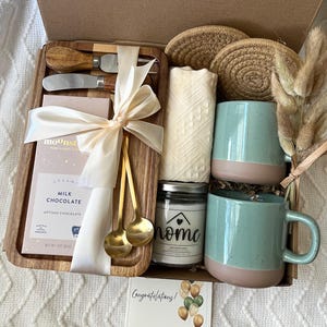 May include: A gift box containing a wooden tray with a milk chocolate bar, two gold spoons, and two cheese knives, all tied with a cream ribbon. Also included are two round woven coasters, a white cloth, a candle labeled home, and two teal mugs.