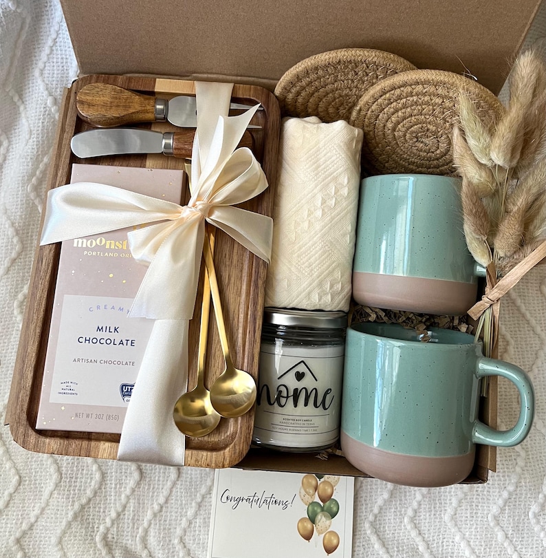 May include: A gift box containing a wooden tray with a milk chocolate bar, two gold spoons, and two cheese knives, all tied with a cream ribbon. Also included are two round woven coasters, a white cloth, a candle labeled home, and two teal mugs.