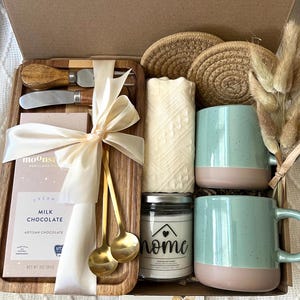 May include: A gift box containing a wooden tray with two gold spoons, a cheese knife, and a 3 oz milk chocolate bar tied with a ribbon. Includes two speckled light blue and tan mugs, a candle labeled home, two woven coasters, and dried grass.