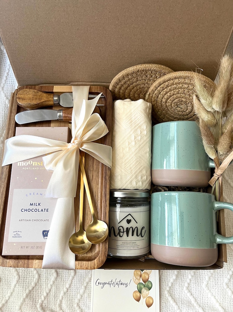 May include: A gift box containing a wooden tray with two gold spoons, a cheese knife, and a 3 oz milk chocolate bar tied with a ribbon. Includes two speckled light blue and tan mugs, a candle labeled home, two woven coasters, and dried grass.