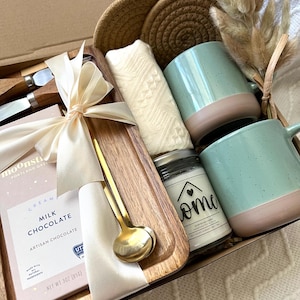 May include: A gift box containing a wooden tray, two speckled mint green and tan mugs, a bar of Moonstruck milk chocolate, a gold spoon, two wooden-handled knives, a white textured cloth, two round woven coasters, and a candle labeled Home.
