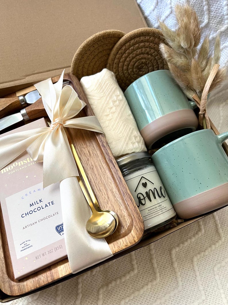 May include: A gift box containing a wooden tray, two speckled mint green and tan mugs, a bar of Moonstruck milk chocolate, a gold spoon, two wooden-handled knives, a white textured cloth, two round woven coasters, and a candle labeled Home.