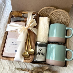 May include: A gift box containing a wooden serving board with two metal spreaders, a bar of Moonstruck milk chocolate, two gold-toned spoons, a cream-colored textile, two round woven coasters, two speckled light blue mugs, and a candle labeled home.