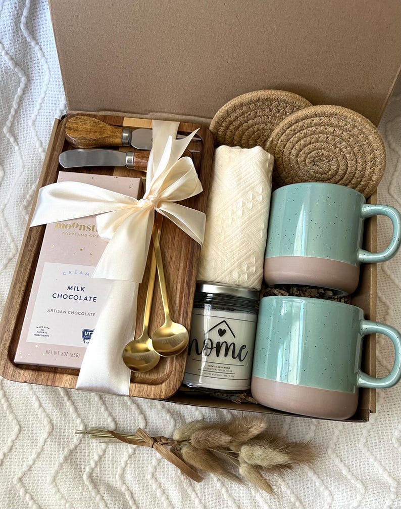 May include: A gift box containing a wooden serving board with two metal spreaders, a bar of Moonstruck milk chocolate, two gold-toned spoons, a cream-colored textile, two round woven coasters, two speckled light blue mugs, and a candle labeled home.