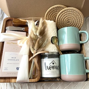 May include: A gift box containing two speckled light green and pink mugs, a glass candle jar labeled home, a wooden tray, two round woven coasters, a 3 oz milk chocolate bar, a gold spoon, two small wooden knives, and a bundle of dried bunny tail grass.