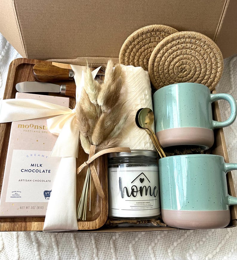 May include: A gift box containing two speckled light green and pink mugs, a glass candle jar labeled home, a wooden tray, two round woven coasters, a 3 oz milk chocolate bar, a gold spoon, two small wooden knives, and a bundle of dried bunny tail grass.
