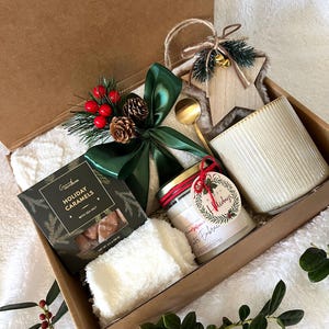 May include: A holiday gift box filled with a candle, caramels, a star-shaped ornament, a small spoon, and a fluffy white towel. The box is decorated with a green bow, pinecones, and red berries. The candle has a red ribbon and a tag that says "Happy Holidays".