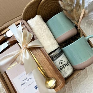 May include: A gift box containing two speckled light blue and pink mugs, a white textured cloth, two round woven coasters, a gold spoon, a wooden tray, two metal utensils with wooden handles, a candle labeled home, and a 3 oz milk chocolate bar.