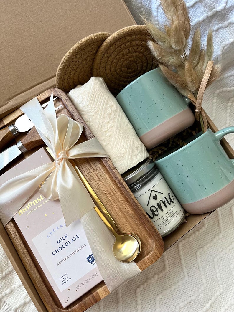 May include: A gift box containing two speckled light blue and pink mugs, a white textured cloth, two round woven coasters, a gold spoon, a wooden tray, two metal utensils with wooden handles, a candle labeled home, and a 3 oz milk chocolate bar.