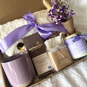 May include: A gift box filled with a lavender-themed assortment. It includes a purple mug, a bar of dark chocolate, a candle, a jar of sweet caramel, a small bouquet of dried flowers, and a white blanket, all tied with a purple ribbon.